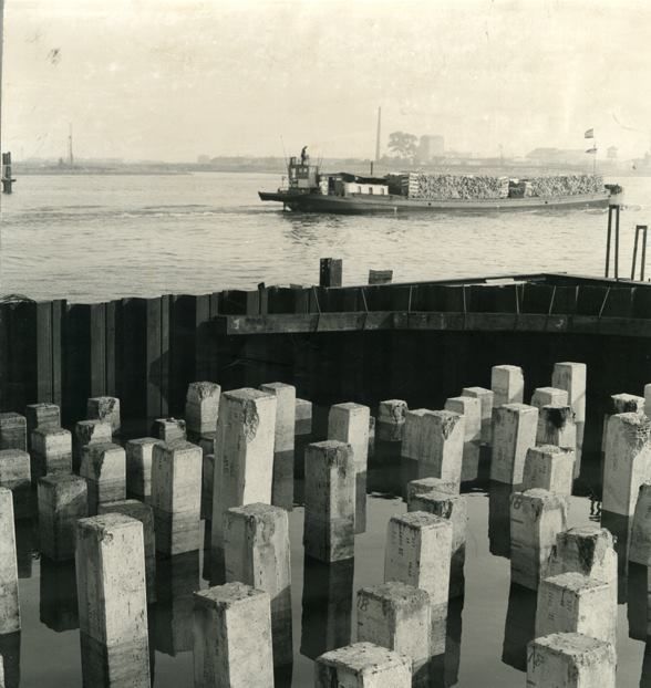 Gorinchem: Als we naar het schip kijken dan lijkt de tijd even stil te staan. Maar schijn bedriegt, want op de voorgrond zien we voorgespannen heipalen van de in aanbouw zijnde Brug over de Merwede. De foto is geschoten in 1959, terwijl de Brug in 1961 door Koningin Juliana werd geopend. Maar ook de binnenvaartschepen zijn inmiddels veranderd. Natuurlijk wordt de Merwede en de Linge nog druk bevaren, maar de goederen staan vaak benedendeks en op de achtersteven zien we vaak een auto staan. Met de komst van de Merwedebrug werd de Alblasserwaard beter ontsloten. Men hoefde niet langer met de auto door de binnenstad om daar het veer op te rijden, met grote wachttijden tot gevolg. Nee, met de auto van Breda naar Utrecht is nu een peulenschilletje..., tenminste als er geen file staat bij bijvoorbeeld het Knooppunt Gorinchem. Maar ook daar wordt aan gewerkt.