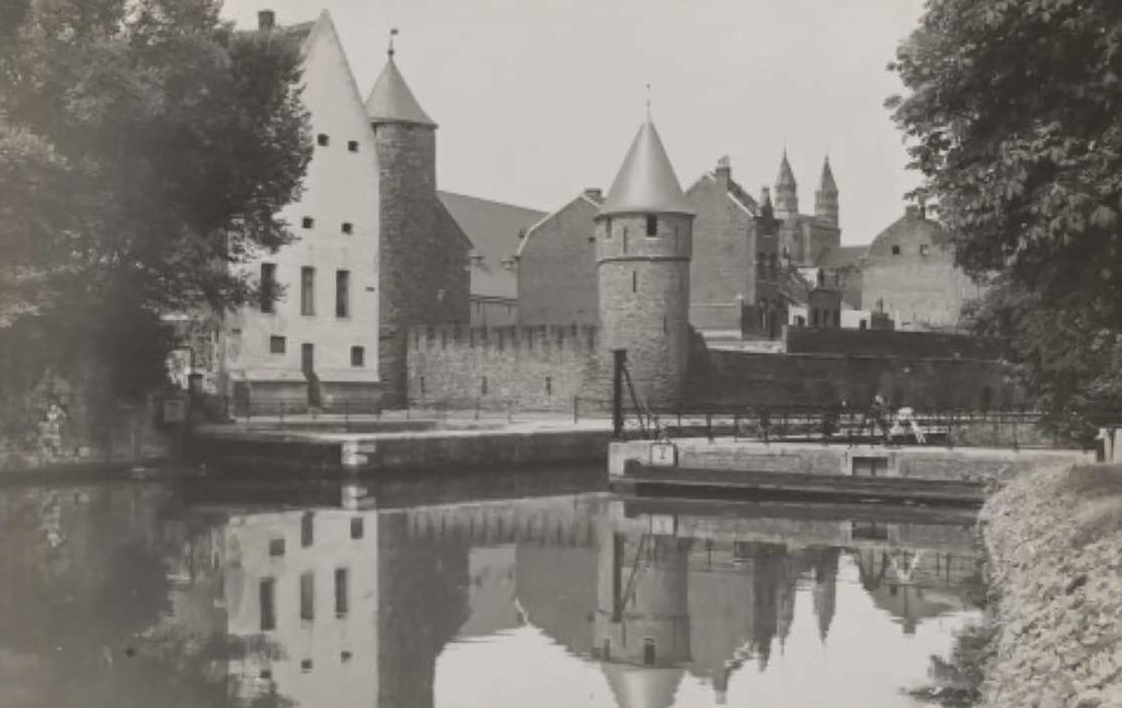 We zien omstreeks 1928 de straten: Vijf Koppen en Helpoort vanaf de overzijde van het in de zestiger jaren gedempte kanaal. Van links naar rechts het Pesthuis, de Helpoort en de Onze Lieve Vrouwewal, met daarboven de torens van het westwerk van de Onze Li