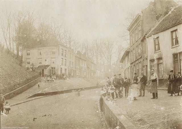 Nijmegen 1895: De Voerweg had vroeger geen beste naam, niet alleen vanwege haar bordelen die met name in de aangrenzende Rozemarijngas aanwezig waren. De man met schort rechtsvooraan op de foto, is Engelbartus Knuvelder, bordeelhouder in de Nijmeegse benedenstad. Hij stond bekend als de grootste pooier van het 19de eeuwse Nijmegen. Links zien we het St. Geertruidsbergje. Op deze originele foto in chamois, kijken we in de richting van het Valkhofplein, midden op de achtergrond (nog net zichtbaar) de Voetgangersbrug, de verbinding tussen het Kelfkensbos en het Valkhof. | De Rozemarijngas dateert uit begin vijftiende eeuw en werd toen ook wel After 't Slot en Achter dat Hoff genoemd. Ze lag aan de voet van de Valkhofheuvel en liep af in de richting van de Waal. Uit 1563 is de naam Rozemaryngas bekend. In de daaropvolgende eeuwen droeg het straatje aan de voet van de Valkhofheuvel ook namen als Villergas, Vildersgas, Goudgraversgas en Stillevegersgas. De straatnamen verwijzen naar de beroepen die door de bewoners van het gasje werden uitgeoefend. Stillevegers, ook wel goudgravers genoemd, waren de voorgangers van de gemeentereiniging. Nijmegen was tot eind negentiende eeuw een vestingstad. De Rozemarijngas was in die tijd vooral berucht om haar bordelen. De straat verkeerde in slechte staat. De woningen waren er totaal vervallen. De bordelen brachten veel overlast met zich mee. Er werd veel geklaagd over het wangedrag van de soldaten. In 1895 werd een begin gemaakt met de sloop van de meest beruchte huizen. Het gasje verdween begin twintigste eeuw van de stadsplattegrond. Mogelijk dankte de gas haar naam aan de prostitutie. Rosemarijnen, ook wel ‘verciersters’ geheten, oefenden hier hun beroep uit. | Het dal waar de Voerweg doorheen loopt is gegraven in de 15e eeuw. Tot die tijd vormden het Valkhof en het Kelfkensbos één geheel. Over de Voerweg konden goederen makkelijker van de Waaloever naar de stad worden vervoerd, omdat deze lange helling minder steil was dan de oorspronkelijke opgang. Tevens vormde de weg een droge gracht die de burcht op het Valkhof beschermde. Aan weerskanten van de weg stonden rijen huisjes. Veel van deze huizen waren al voor 1900 afgebroken. Aan het einde van de Voerweg stond de Hunner- of Hoenderpoort. Deze is gesloopt aan het einde van de negentiende eeuw met de ontmanteling van de vestingstad.
