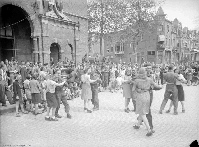 Nijmegen 1945: Deze foto toont de uitzinnige Nijmeegse bevolking op 5 mei 1945. Het is Bevrijdingsdag. Mensen dansen op het Hertogplein voor de Nutsschool. Links zicht op de Gerard Noodtstraat en op de Van der Brugghenstraat. | Alhoewel Nijmegen al in september 1944 bevrijd was door de Geallieerden bleef de stad tot het oorlogseinde een frontstad en dat betekende dat het oorlogsgevaar nog lang niet geweken was. De al door het bombardement van 22 februari 1944 geteisterde stad werd zowel in het centrum als daarbuiten nog meer gehavend. Weer waren er burgerslachtoffers te betreuren. Dagenlang was er verschrikkelijk geschoten. Overal lagen doden en gewonden. Op het Mariënburg ontplofte een munitiewagen. Wegtrekkende Duitsers en leden van de Hitlerjugend staken in de stad gebouwen en woonhuizen in brand. Zeven maanden ligt de stad onder vuur, met ingrijpende gevolgen: honderden doden en gewonden en een verwoest centrum. De burgerij verbleef dag en nacht in schuilkelders, levend tussen hoop en vrees. | Nijmegen verandert in die periode in één groot militair kampement: de springplank voor het grote Rijnland-offensief dat in het voorjaar van 1945 eindelijk de Duitse weerstand breekt. In die periode zijn de contacten tussen Nijmeegse burgers en de buitenlandse militairen zeer intensief.