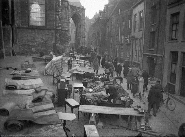 Nijmegen 1937: St. Stevenskerkhof, de 'Lusemert' | Grabbelen en graaien tussen oude, tweedehands spullen. De Lusemert in Nijmegen is in de loop der jaren maar weinig veranderd. Sommige mensen komen er langer dan vijftig jaar. De Lusemert (luizenmarkt) in Nijmegen is een begrip. In vroeger tijden was de oude Benedenstad een enorme opslagplaats van lompen. Het is daarom niet verwonderlijk dat elke maandagmorgen aan de voet van de St. Stevenstoren de Lusemert gehouden werd. Een schilderachtige maar toch ook wel armelijke markt in tweedehands goederen.