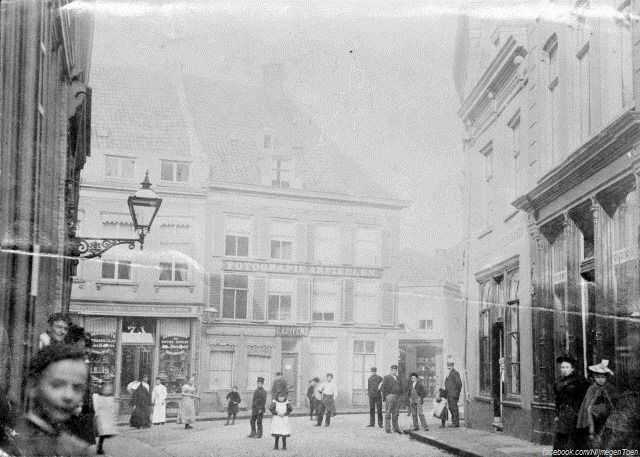 Nijmegen 1895: Op dit prachtige tijdsbeeld kijken we in het oude Nijmegen van de 19e eeuw vanuit de Ganzenheuvel in de richting van de Priemstraat. We zien de panden van fotograaf C.A.P. Ivens en drogist H.A. Tesser. Links is de Lange Brouwerstraat.