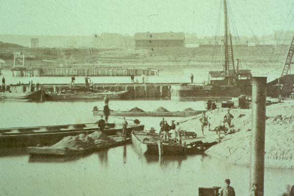 Aanleg van de spoorbrug over de Waal, gezien vanaf de Lentse oever. Op de achtergrond fabrieken aan de Nieuwe Haven. Foto uit circa 1875.