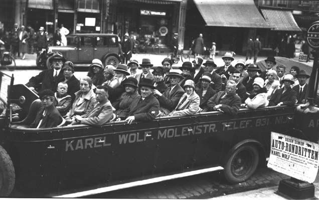 Nijmegen: Een omnibus in de Molenstraat, waar de reizigers aan het begin staan van een toertje rond Nijmegen. Ze poseren nogal stijfjes voor een professionele fotograaf die van de gelegenheid gebruik maakt om zijn nering te vergroten. Na afloop kunnen de toeristen, verrukt over zoveel moois in de omgeving, de foto als souvenir aanschaffen. Het lijkt Disneyland wel! De foto dateert uit 27 juli 1929.