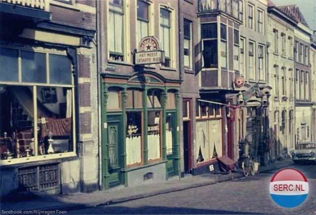 Nijmegen: De Priemstraat 1960. We kijken in de richting van de Lage Markt met de karakteristieke groene houten gevel van Café A. ten Oever en de naastgelegen Appie Bar