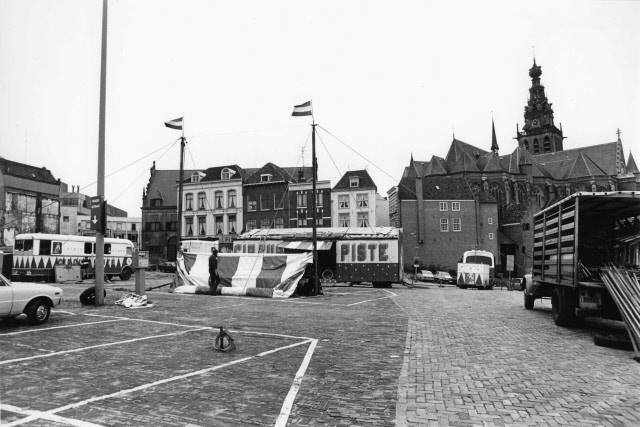 De opbouw van het circus 'Piste' op een kale Korenmarkt in 1977. De Korenmarkt werd na en tijdens de afbraak van een deel van de Benedenstad gebruikt als tijdelijke parkeerplaats zoals zoveel plekken in die tijd. Een ideale plek om een circustent neer te zetten. Het geeft een mooie situatiefoto van hoe het er uitzag aan het eind van de jaren '70. We zien de achterzijde van de Waag, de nog steeds bestaande huizenrij aan de Kannenmarkt en natuurlijk de St. Stevenskerk.