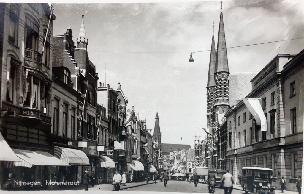 De Molenstraat omstreeks 1930. We zien de neo-gotische Canisiuskerk met haar kathedrale pronkgevel en ranke westtorens. Op de achtergrond zien we de torens van de Broerstraatkerk. Het pand rechts op de foto is het Oud Burgeren Gasthuis (nu is hier de ingang van de Molenoort passage). Voordat het gebouw tijdelijk hoofdbureau van politie werd en daarna onder de slopershamer zijn einde vond, waren hier in 1930 nog de oude burgers van Nijmegen gehuisvest. Buiten de pastorie is van dit gehele huizenblok, vanaf de Ziekerstraat tot en met het perceel rechts van het gasthuis, niets meer over gebleven. De Molenstraat is altijd al een echte winkelstraat geweest. Veel van de panden aan de overzijde van de kerk staan er nog steeds.