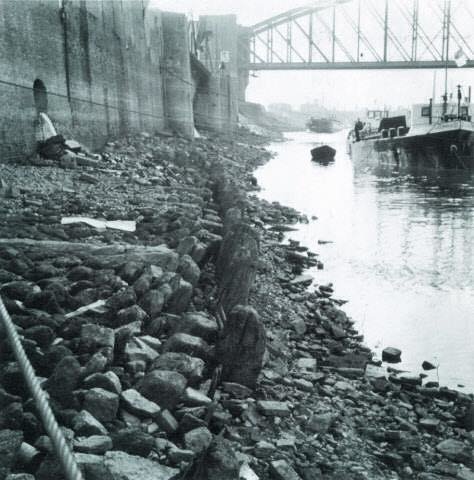In 1953 zag de kade er heel anders uit dan tegenwoordig het geval is. We zien een robuuste hoge kademuur en het riool dat nog vrij in de Waal kon lozen. Op de achtergrond de oude Spoorbrug.