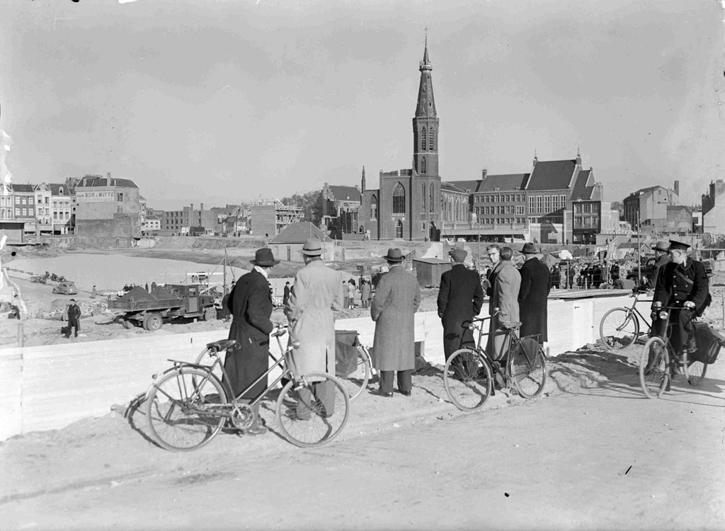In hartje stad kijkt de Nijmeegse bevolking in 1948 toe bij de werkzaamheden voor de aanleg van het splinternieuwe Plein 1944. Het nieuwe plein gaf de stad tijdens de wederopbouw een heel nieuw gezicht. In het midden de verdwenen Dominicuskerk (ook wel bekend als de Broerstraatkerk) en rechts daarvan het Stadhuis. Op de achtergrond links, de achterzijde van kledingzaak van Van Dijk & Witte aan de Grote Markt.