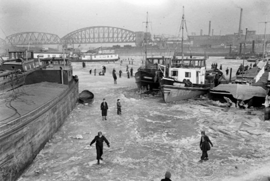De dichtgevroren haven in 1954. Mensen wandelen op het ijs. Op de achtergrond de Waal-Spoorbrug.