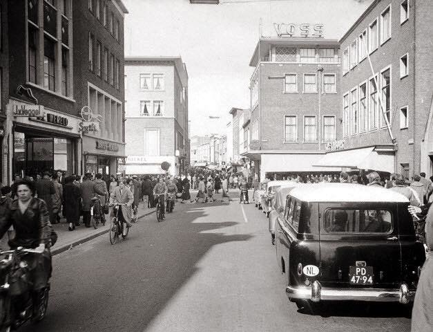 de Molenstraat in Nijmegen omstreeks 1960. Vanaf de Molenstraat kijken we in de richting van de kruising met Plein 1944, Broerstraat en de Ziekerstraat. Een druk kruispunt is het altijd al geweest. Op drukke winkelwagen zag je regelmatig een verkeersagent op een sokkel staan op dit punt. Links op de foto zien we de winkel van Van Veggel Schoenmode (sinds 1919!) en daar achter de zelfbedieningswinkel in levensmiddelen van Jansen-Hendriks. Op de achtergrond het pand van Modehuis Voss en rechts Kreymborg.