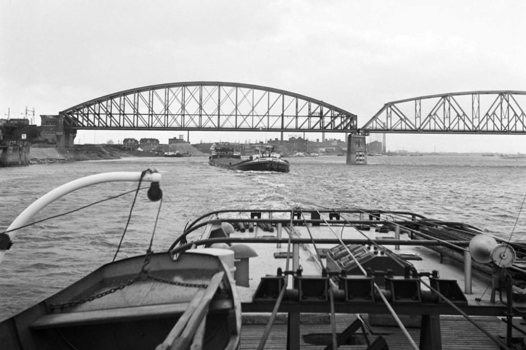 Klik op de foto om alle oude foto's te bekijken - De motorsleepboot DAMCO 9 met zeven schepen in aanhang passeert in de opvaart de spoorbrug van Nijmegen. Op de eerste lengte hangt de afgeladen 3000 ton grote AVONTUUR 6. Foto dateert van voorjaar 1955
